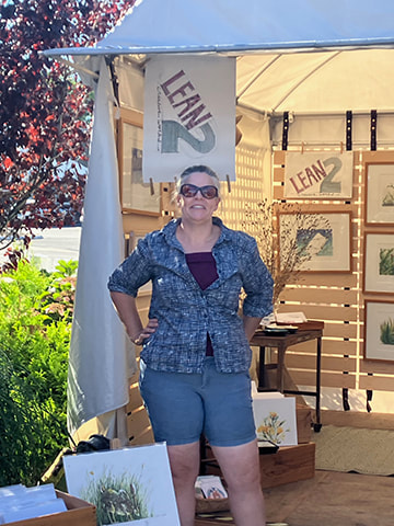 Photo of artist in a blue shirt and shorts in front of a display booth in an outdoor setting.
